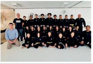 foto de jovens com quimonos pretos de jiu-jitsu, e um homem de a paisana, calça caqui e blusa azul clara, em um dojo branco